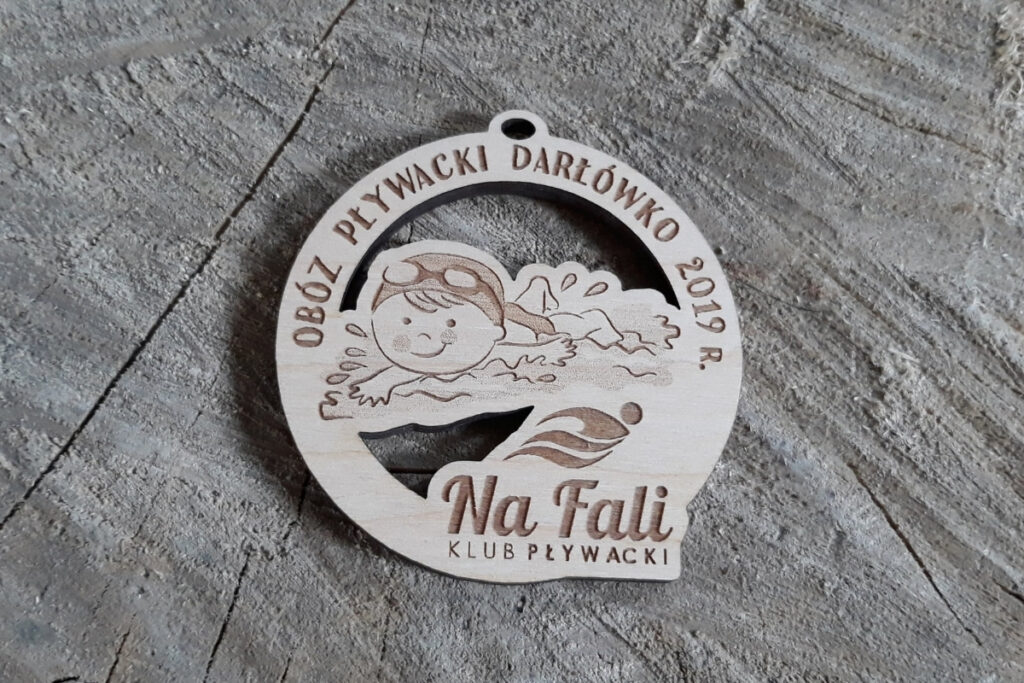 Okrągły drewniany medal z grawerem płynącego dziecka w goglach, napisem Obóz Pływacki Darłówko