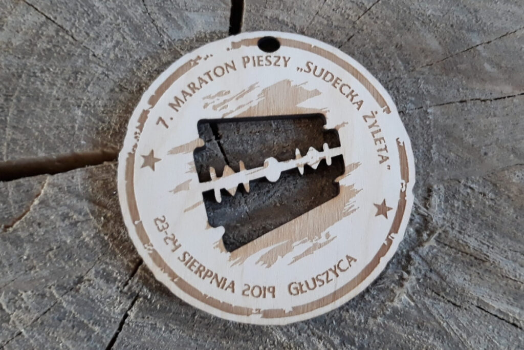 Okrągły drewniany medal z ażurowym wycięciem w kształcie żyletki pośrodku, grawerem ozdobnym i napisem Maraton Pieszy.