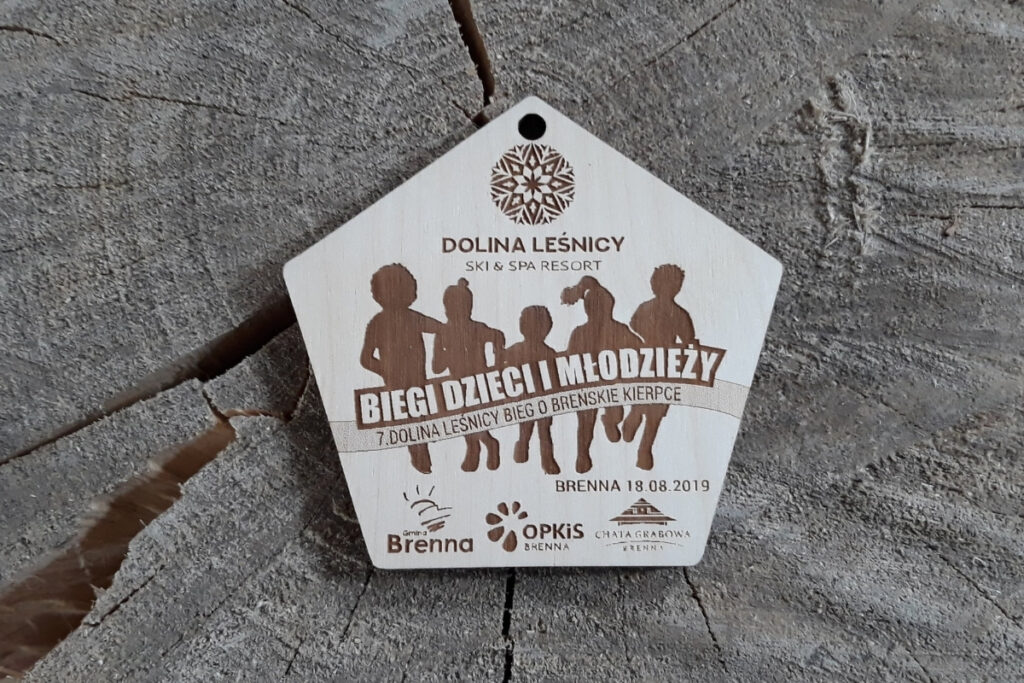 Drewniany medal z grawerem piątki biegnących dzieci w różnym wieku, napisem Bieg Dzieci i Młodzieży oraz logotypami.