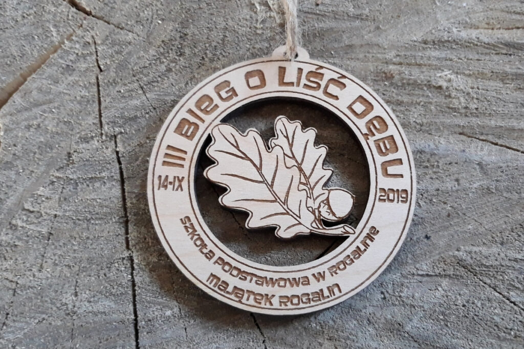 Okrągły drewniany medal z ażurowym, pustym wycięciem w kształcie liścia dębu i grawerem nazwy biegu.