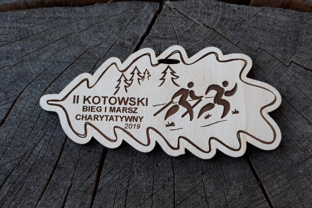 Drewniany medal wycięty w kształcie liścia z grawerem logotypu II Kotowskiego Biegu i Marszu oraz napisami i datą.