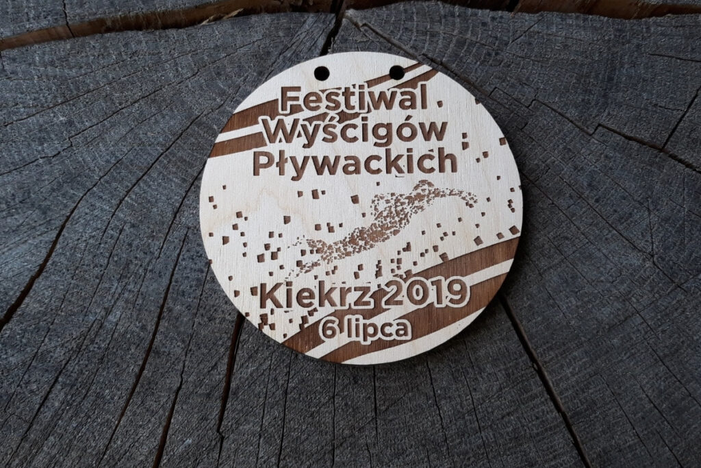 Okrągły drewniany medal z grawerem pływaka w czepku i okularach, napisem Festiwal Wyścigów Pływackich oraz motywem fal.