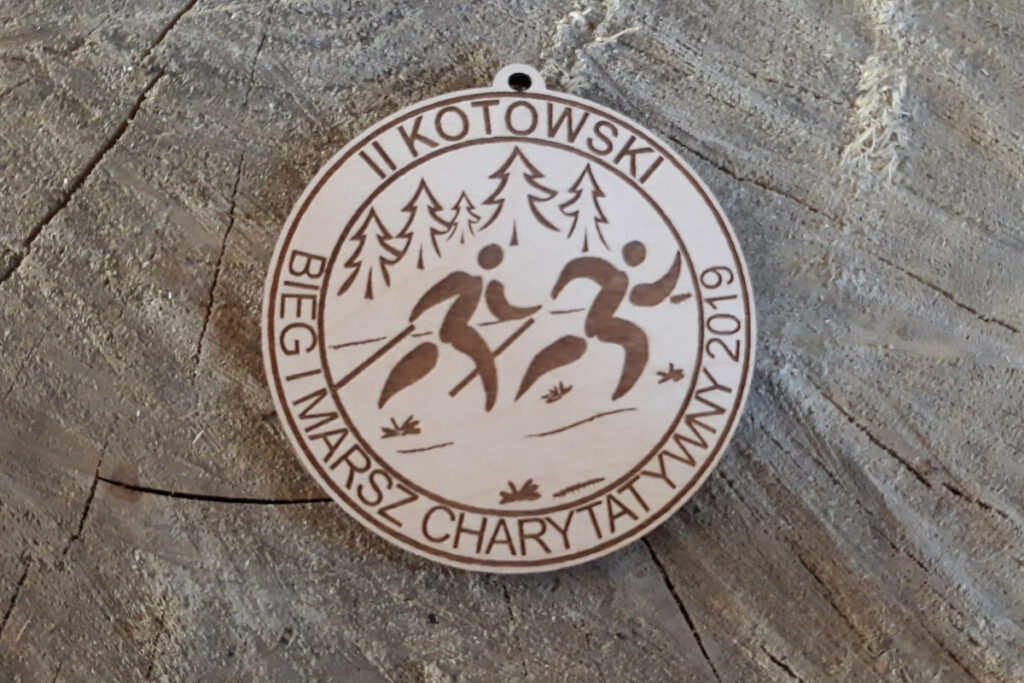 Okrągły drewniany medal z grawerem logotypu z kotem, napisem II Kotowski Bieg i Marsz Charytatywny oraz hasłem Pomagamy.