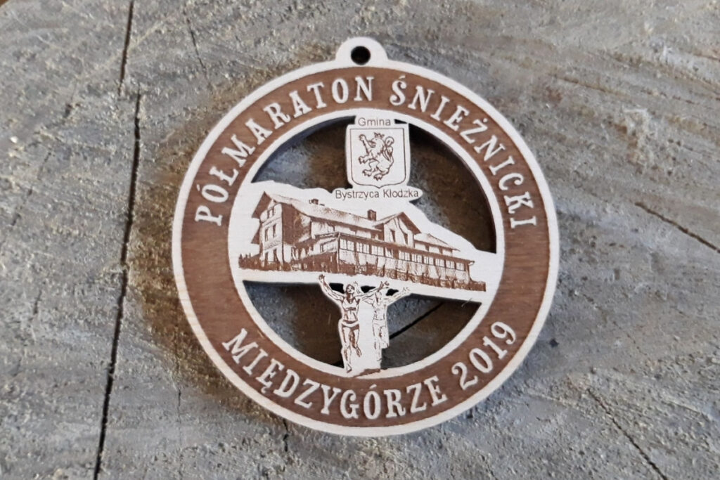 Okrągły drewniany medal z ażurowo wyciętym konturem góry Śnieżnik, sylwetką biegacza i grawerowanym napisem Półmaraton Śnieżnicki.