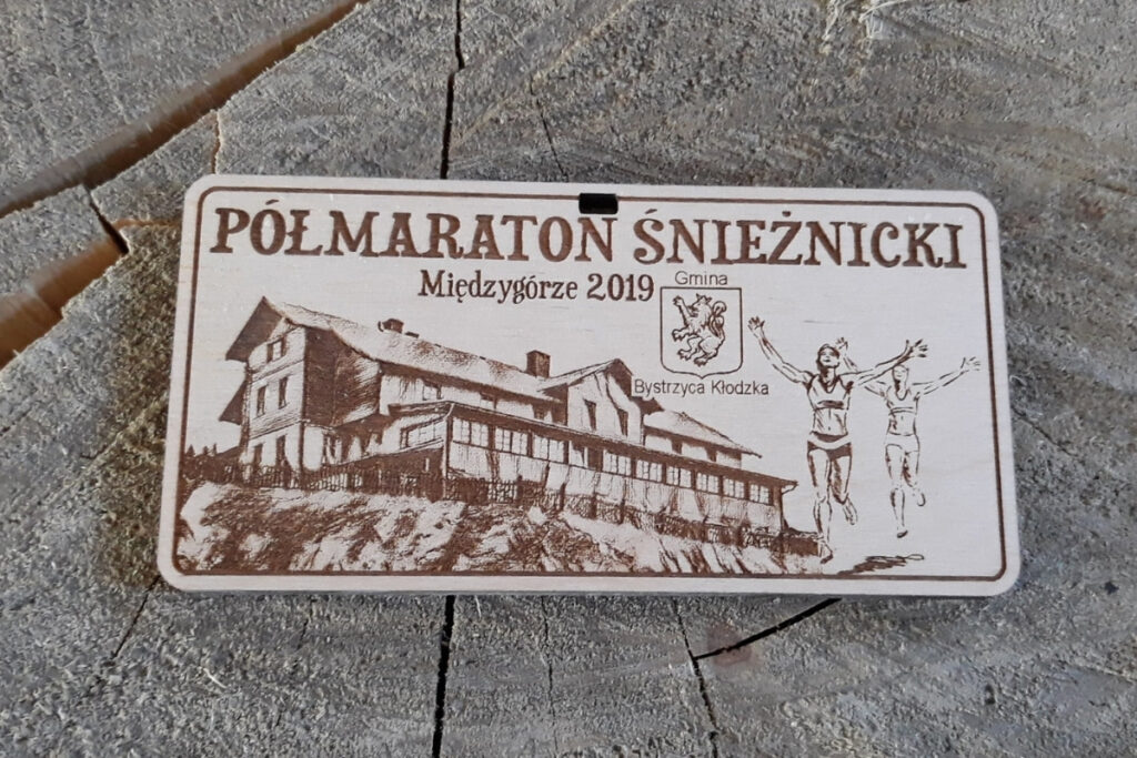 Prostokątny drewniany medal z grawerem przedstawiającym schronisko na Śnieżniku, panoramę gór oraz sylwetkę biegacza.
