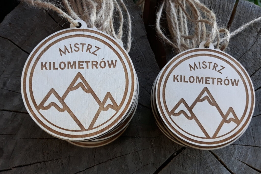 Okrągły drewniany medal z grawerem uskrzydlonego buta biegowego, wieńcem laurowym i napisem Mistrz Kilometrów.