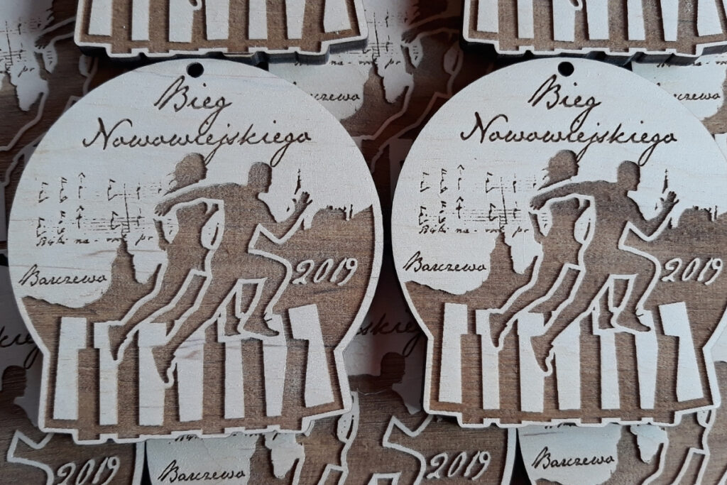 Okrągły drewniany medal z grawerowanym portretem Feliksa Nowowiejskiego, napisem Bieg Nowowiejskiego oraz motywem pięciolinii i nut.