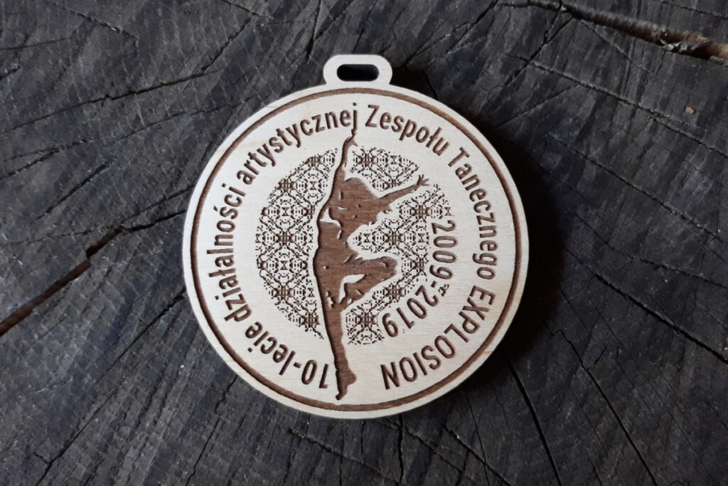 Okrągły drewniany medal z grawerem logotypu zespołu artystycznego, napisem 10-lecie oraz tancerką.