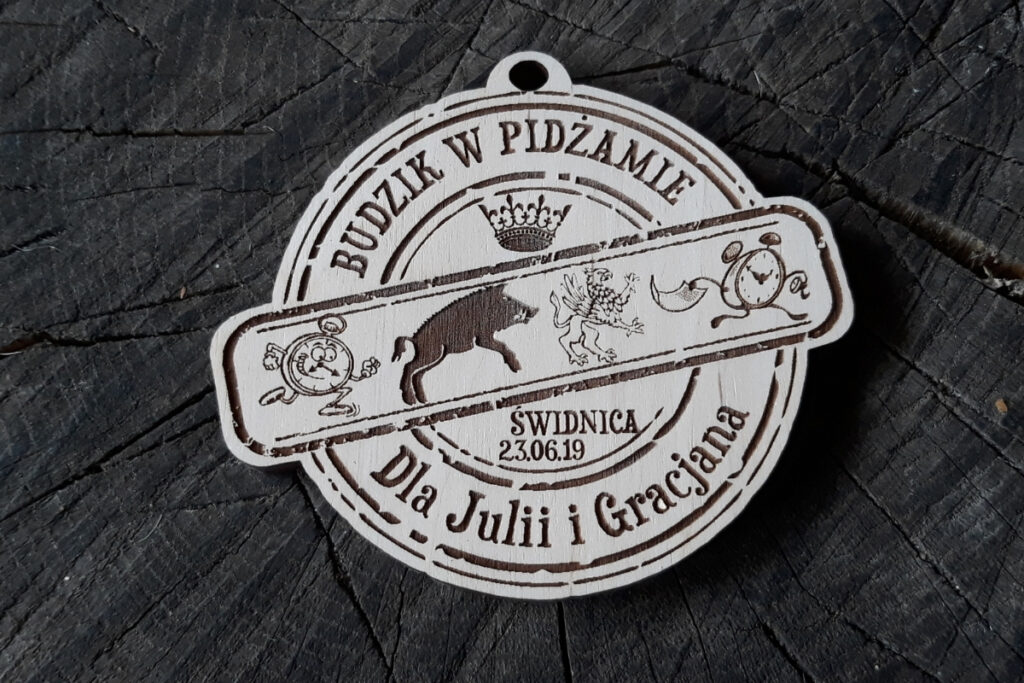 Okrągły drewniany medal z grawerem budzika, napisem Bieg Budzika dla Julii i Gracjana oraz datą wydarzenia.
