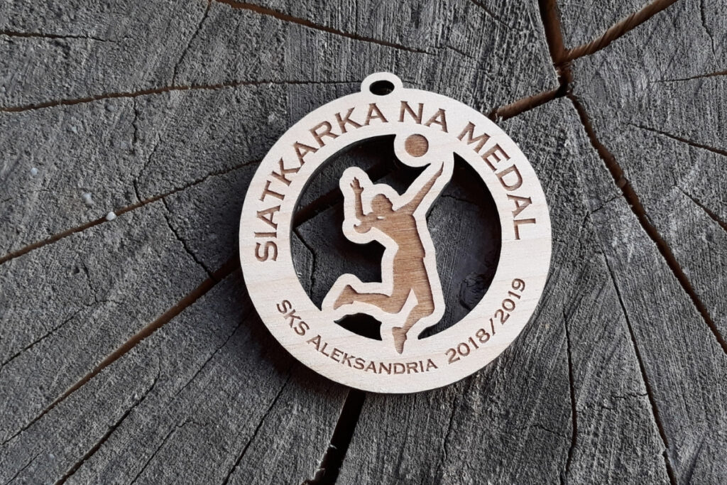 Okrągły drewniany medal z ażurowo wyciętą sylwetką siatkarki w wyskoku do ścinania piłki oraz grawerem nazwy wydarzenia.