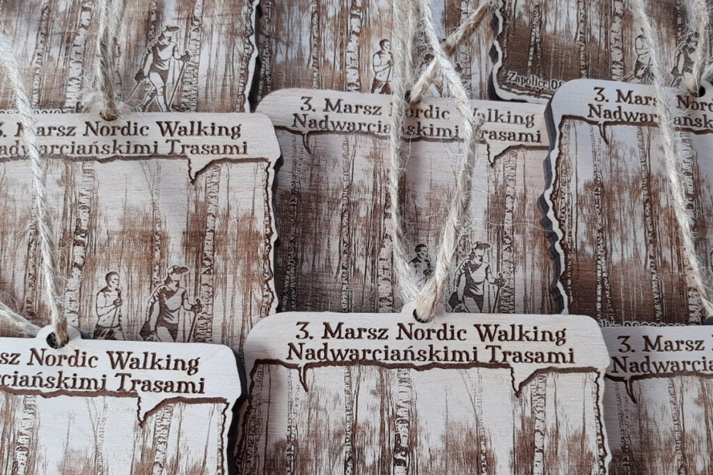 Drewniane medae z grawerem dwóch osób uprawiających Nordic Walking na tle gęstego lasu brzozowego, medale prostokątne, na sznurkach.