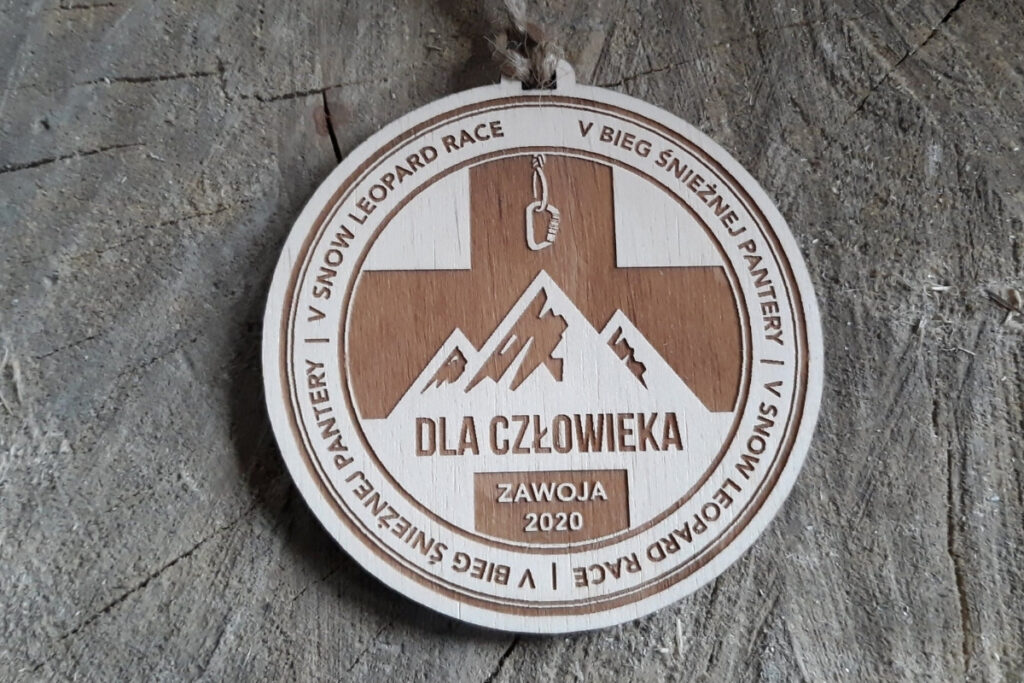 Okrągły drewniany medal z grawerem  ostrych górskich szczytów, napisem V Bieg Śnieżnej Pantery oraz logotypem wydarzenia.