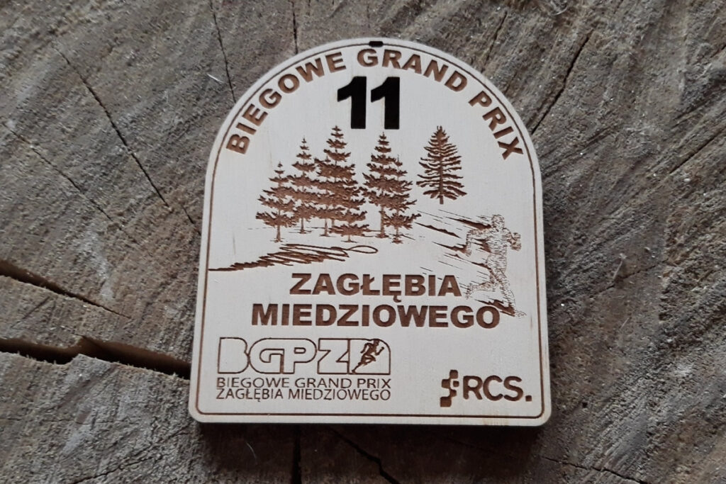 Drewniany medal na 11. Biegowe Grand Prix Zagłębia Miedziowego, z grawerem logotypu cyklu, motywem biegacza i partnerów, wykonany z jasnej brzozy z czystym grawerem bez przypaleń.