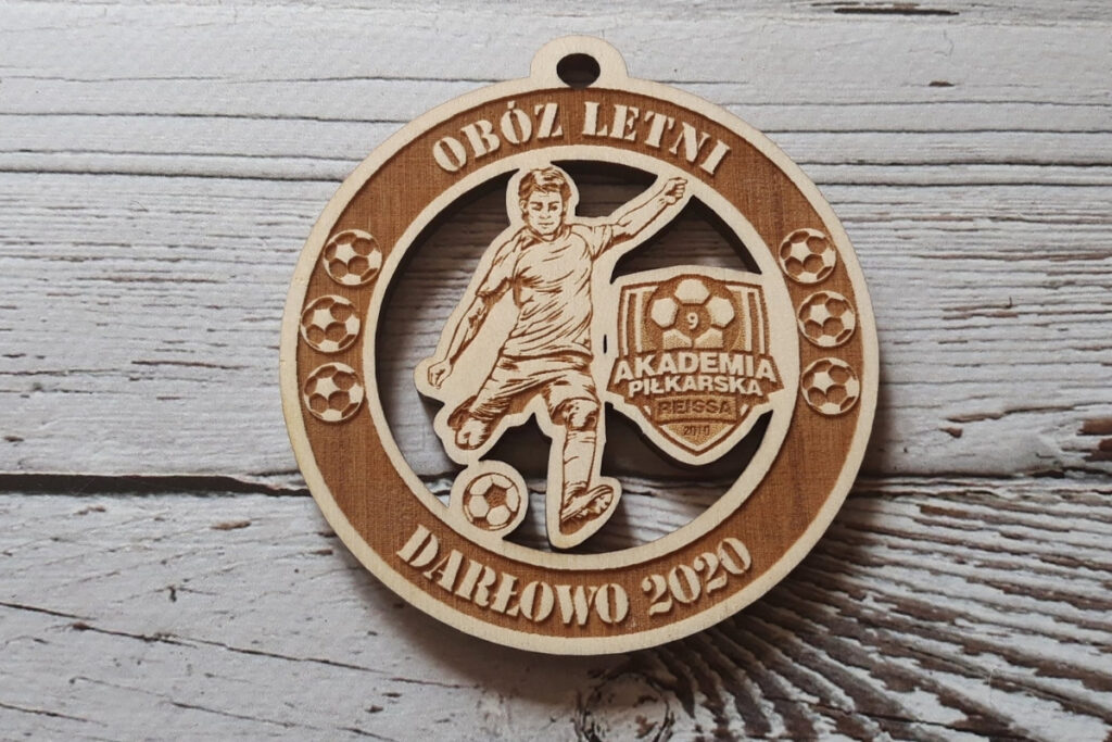 Okrągły drewniany medal ażurowy z wyciętą sylwetką piłkarza prowadzącego piłkę, personalizowany grawer na obóz letni, wykonany z jasnej brzozy bez śladów przypaleń.