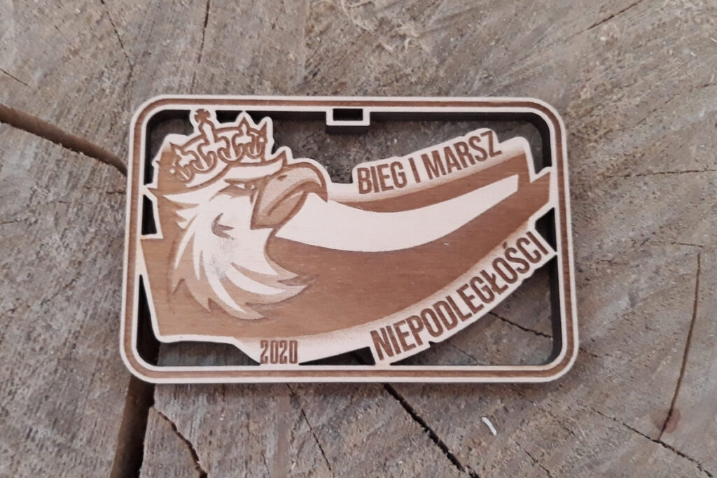 Prostokątny drewniany medal patriotyczny na Bieg i Marsz Niepodległości, z ażurowo wyciętym orłem w koronie, grawerem biało-czerwonej flagi oraz datą 11 listopada, wykonany z jasnej brzozy bez osmaleń.