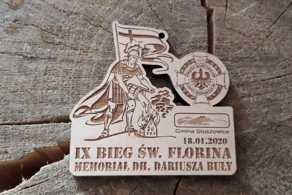 Drewniany medal na IX Bieg św. Floriana z grawerem postaci świętego gaszącego pożar, logotypem wydarzenia oraz ozdobną typografią, wykonany z jasnej brzozy bez śladów osmaleń.