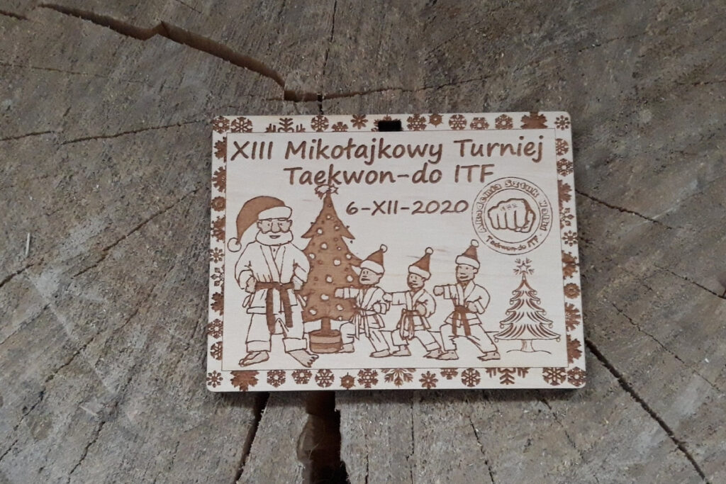 Prostokątny drewniany medal na XIII Mikołajkowy Turniej Taekwon-do ITF z grawerem zawodnika wykonującego kopnięcie boczne, czapką św. Mikołaja i logotypem federacji, czyste wykończenie bez osmaleń.