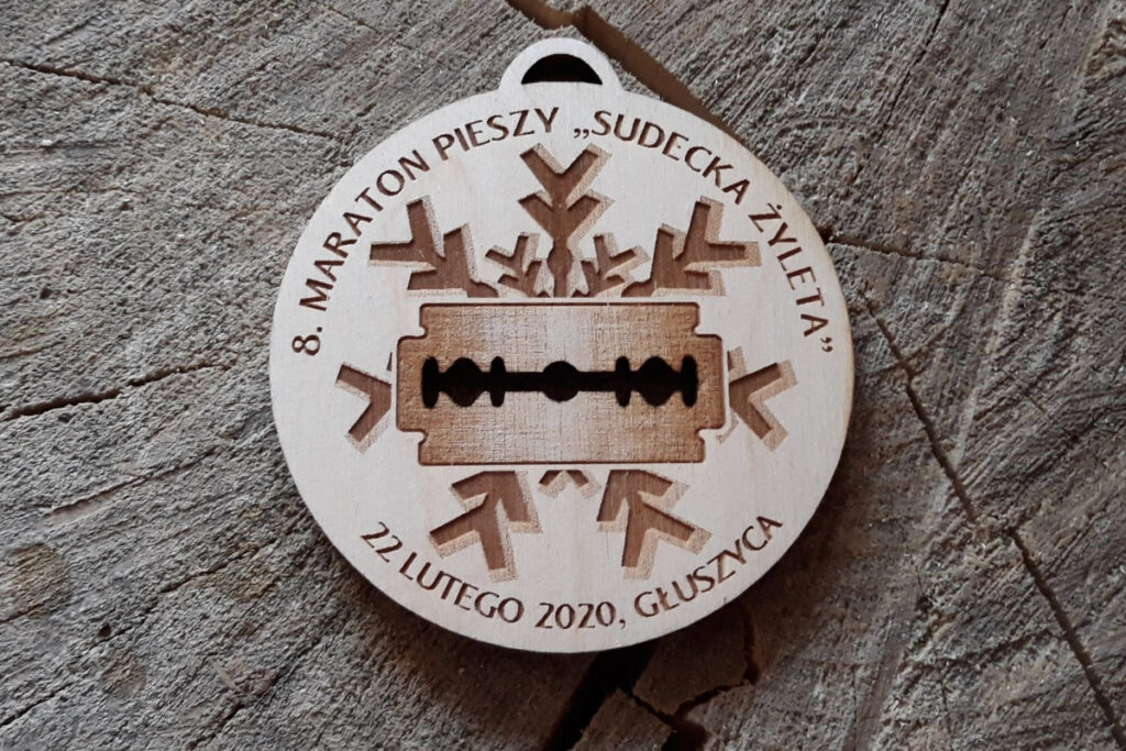 
Okrągły drewniany medal na 8. Maraton Pieszy Sudecka Żyleta, z ażurowo wyciętym motywem żyletki w środkowej części oraz grawerem płatka wykonany z jasnej brzozy bez osmaleń.