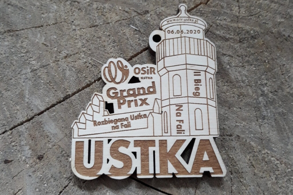 Drewniany medal na Grand Prix Ustki z precyzyjnym grawerem zabytkowej latarni morskiej, logotypem wydarzenia oraz ozdobną falą, wykonany z jasnej brzozy bez osmaleń.