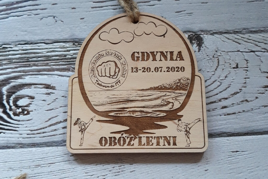 Półokrągły drewniany medal pamiątkowy z obozu letniego w Gdyni, z grawerem fal i brzegu,  wykonany z jasnej brzozy bez śladów osmaleń.