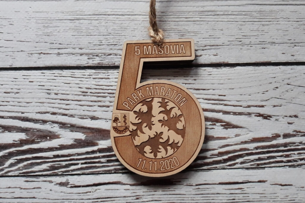 Personalizowany drewniany medal w kształcie cyfry 5, z grawerem logotypu wydarzenia oraz daty, wykonany z jasnej brzozy bez śladów przypaleń, zawieszony na kolorowej tasiemce.