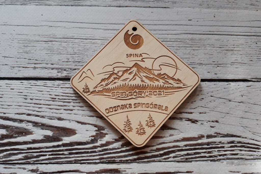 Okrągły drewniany medal „Odznaka Śpingórala” z grawerem górskich szczytów, oraz stylizowaną nazwą wydarzenia, wykonany z czystej brzozy bez śladów przypaleń.
