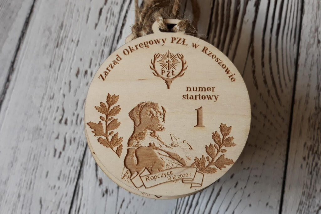 Drewniany medal - tabliczka z numerem startowym na zawody Polskiego Związku Łowieckiego, z grawerem logotypu PZL, numerem zawodnika oraz ozdobnymi motywami roślinnymi, wykonana z jasnego drewna bez osmaleń.