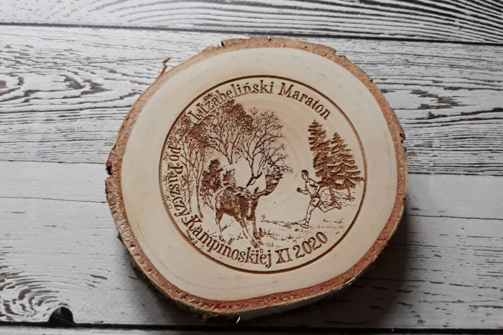 Drewniany medal na Maraton po Puszczy Kampinoskiej, wykonany na naturalnym plastrze brzozy z korą, z grawerem łosia, logotypem wydarzenia oraz datą, czysta powierzchnia bez osmaleń.