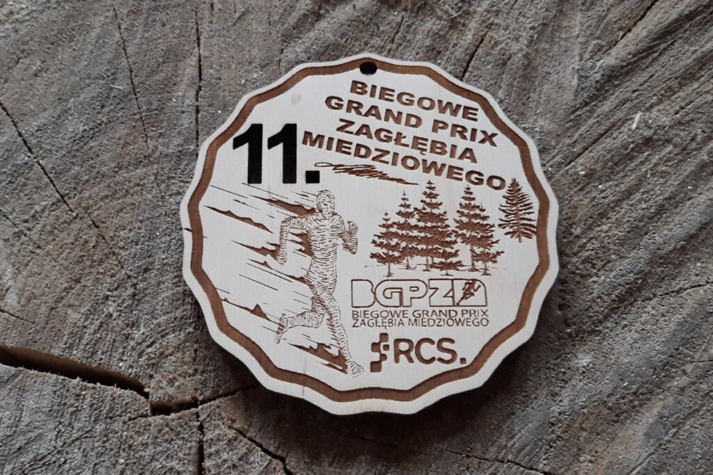 Okrągły drewniany medal na 11. Biegowe Grand Prix Zagłębia Miedziowego, z grawerem logotypu wydarzenia, numerem edycji oraz wieńcem biegaczem pośród drzew wykonany z jasnej brzozy bez śladów dymu.