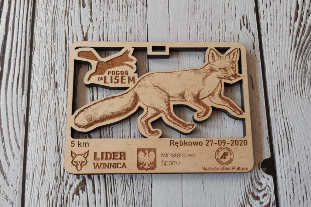 Prostokątny drewniany medal na bieg „Pogoń za lisem”, z ażurowo wyciętą sylwetką biegnącego lisa oraz grawerem logotypu wydarzenia, wykonany z płyty z jasnej brzozy bez śladów dymu.
