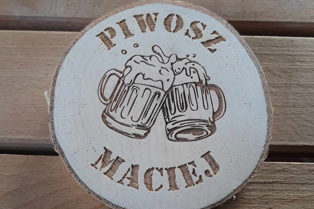 Okrągła drewniana podstawka pod piwo z grawerem „Piwosz Maciej”, grafiką kufla z pianą oraz ozdobnymi wykonana z jasnego plastra brzozy bez śladów osmaleń.