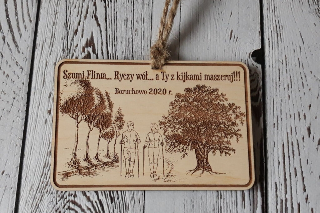 Prostokątny drewniany medal z humorystycznym napisem „Szumi Flinta… Ryczy Wół… a Ty z kijkami Maszeruj!!!”, grafiką rzeki, lasu i kijków do Nordic Walking, wykonany z czystej brzozy bez osmaleń.