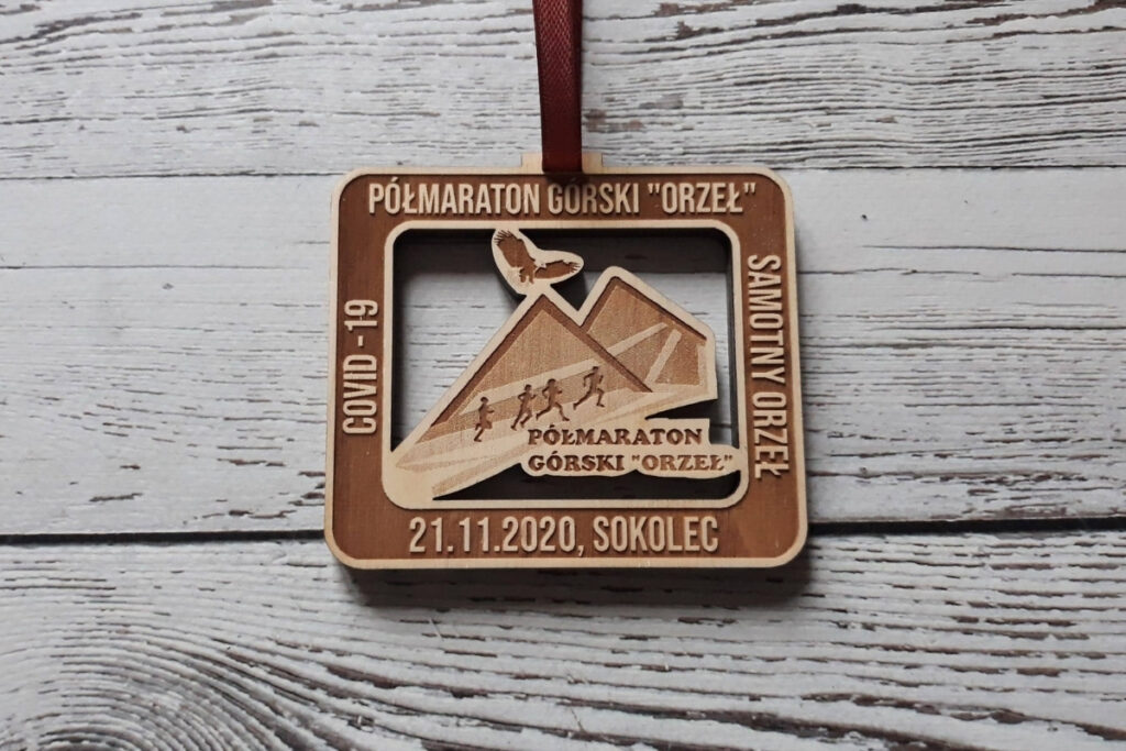 Kwadratowy drewniany medal na Półmaraton Górski z ażurowo wyciętą panoramą szczytów, grawerem biegaczy na szlaku oraz ptakiem nad szczytami, wykonany z jasnej płyty brzozy bez śladów dymu i przypaleń, głęboko wygrawerowany.