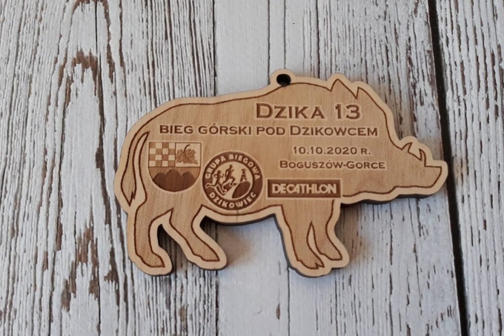Drewniany medal o nieregularnym kształcie postury dzika, z logotypem wydarzenia, wykonany z jasnej brzozy bez śladów dymu i przypaleń.
