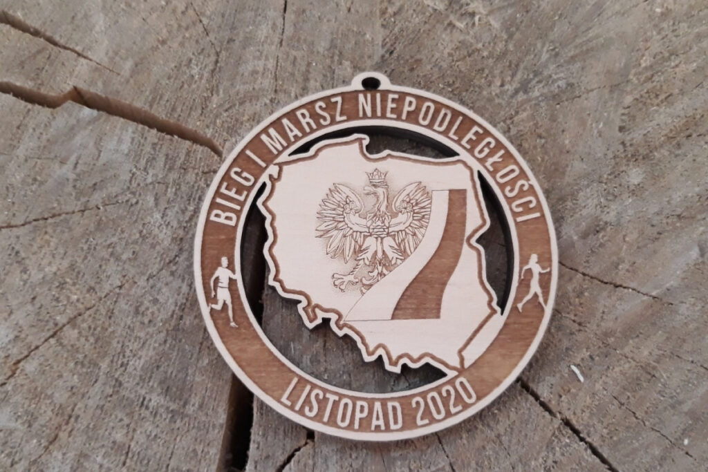 Okrągły drewniany medal na Bieg Niepodległości, z precyzyjnie wyciętym ażurowym konturem Polski wewnątrz, grawerem daty 11 listopada oraz napisami, Orłem i flagą, wykonany z jasnej płyty brzozy bez śladów osmaleń.
