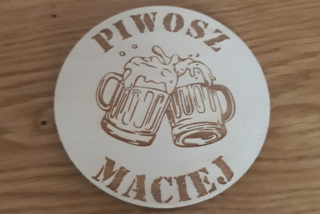 Okrągła drewniana podstawka pod kufel z grawerem „Piwosz Maciej”, grafiką pienistego kufla piwa, wykonana z jasnej sklejki brzozowej bez śladów dymu i osmaleń.