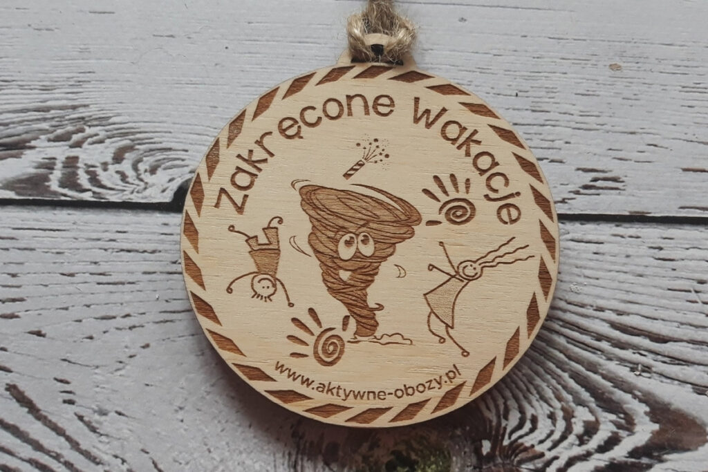 Okrągły drewniany medal pamiątkowy z napisem „Zakręcone Wakacje”, radosną grafiką letniego słońca, uśmiechu oraz elementami wakacyjnej przygody, wykonany z jasnej brzozy bez przypaleń i osmaleń.