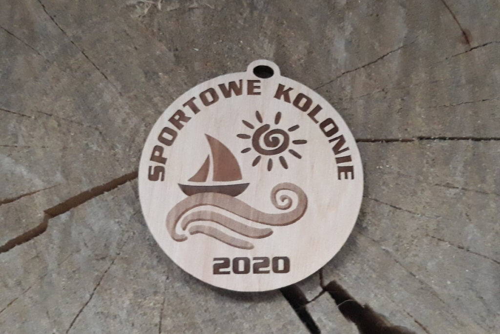 Tekst alternatywny (Alt Text):
Okrągły drewniany medal pamiątkowy na „Sportowe Kolonie”, z grawerem żaglówki płynącej po falach pod uśmiechniętym słońcem, wykonany z czystej brzozy bez śladów przypaleń i osmaleń.