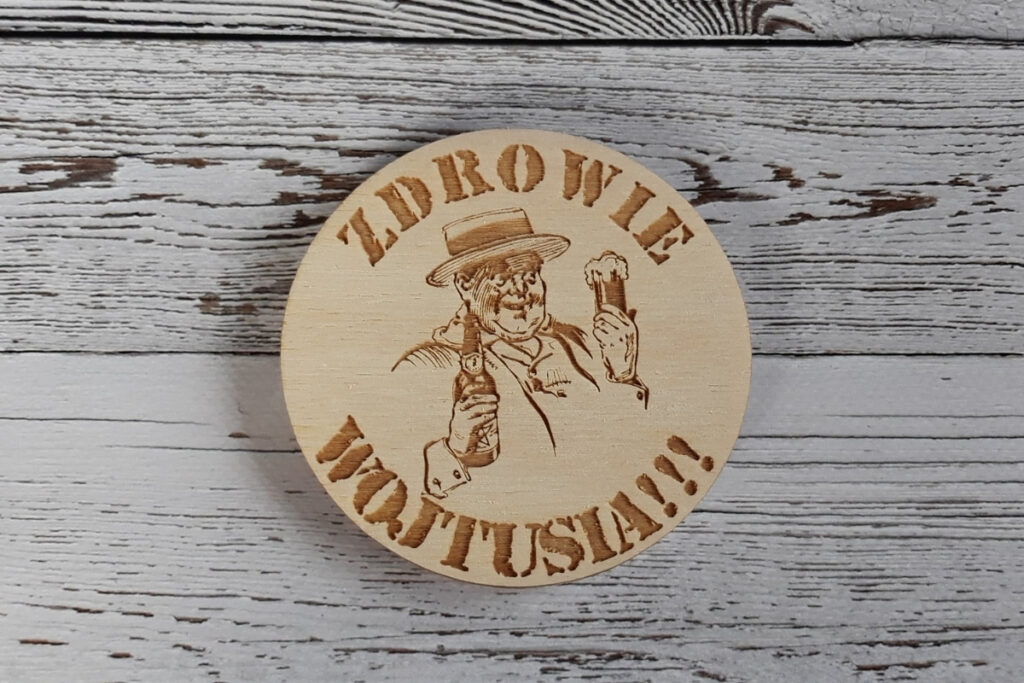 Okrągła drewniana podstawka pod piwo z grawerem „Zdrowie Wojtusia”, grafiką kufla z pianą w rękach pijaka, wykonana z jasnej sklejki brzozowej bez śladów osmaleń i przypaleń.