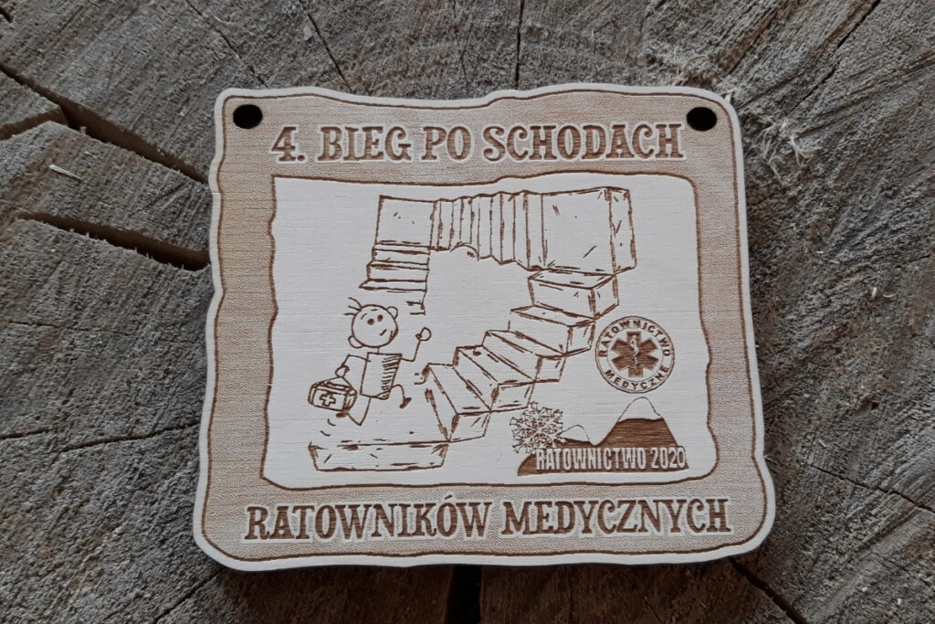 Prostokątny drewniany medal na „Bieg po schodach” dla Ratowników Medycznych, z grawerem Gwiazdy Życia (symbolu ratownictwa), sylwetką biegacza oraz nazwą zawodów, wykonany z jasnej brzozy bez śladów dymu i osmaleń. Biegacz biegnie po schodach z torbą medyczną. Obok logo Ratownictwo 2020.