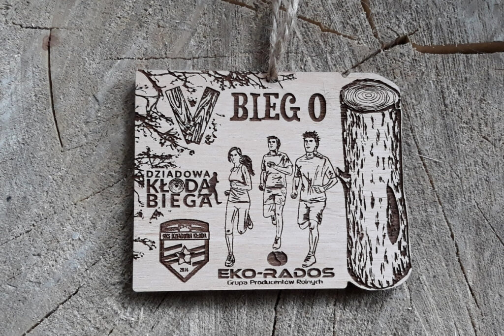 Drewniany medal na „V Bieg o Kłodę”, z grawerem stylizowanej kłody drewna, liści w rogu oraz numerem edycji i biegaczami, wykonany z czystej płyty brzozy bez śladów przypaleń, z dopasowanym naturalnym sznurkiem jutowym.
