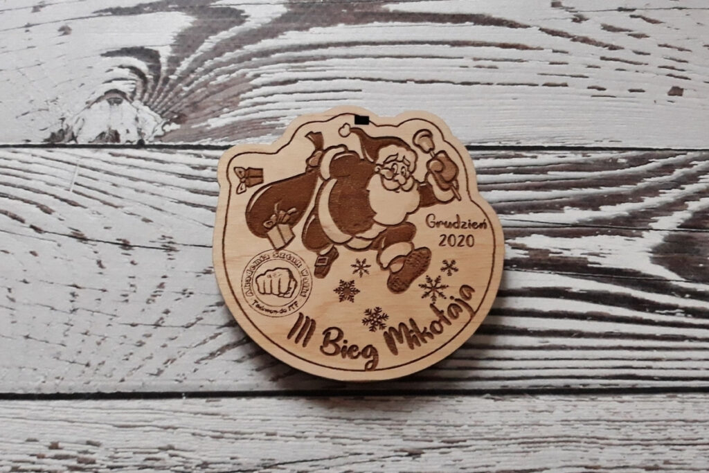 Drewniany medal na „III Bieg Mikołaja”, z radosnym grawerem biegnącego Świętego Mikołaja w czapce z pomponem, otoczony motywami płatków śniegu i nazwą zawodów, wykonany z jasnej brzozy bez przypaleń.