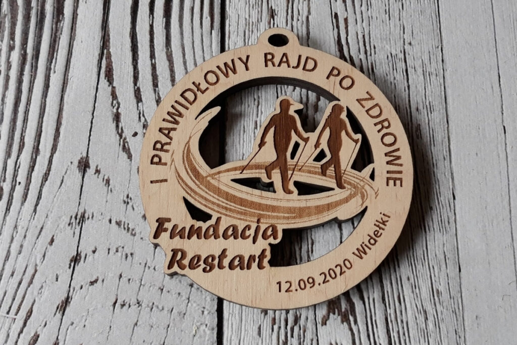 Okrągły drewniany medal na „Rajd Nordic Walking” z precyzyjnym ażurowym wycięciem sylwetek maszerujących osób z kijami oraz grawerem nazwy wydarzenia, nazwy Fundacji - logotypem, wykonany z czystej brzozy bez śladów dymu i osmaleń.