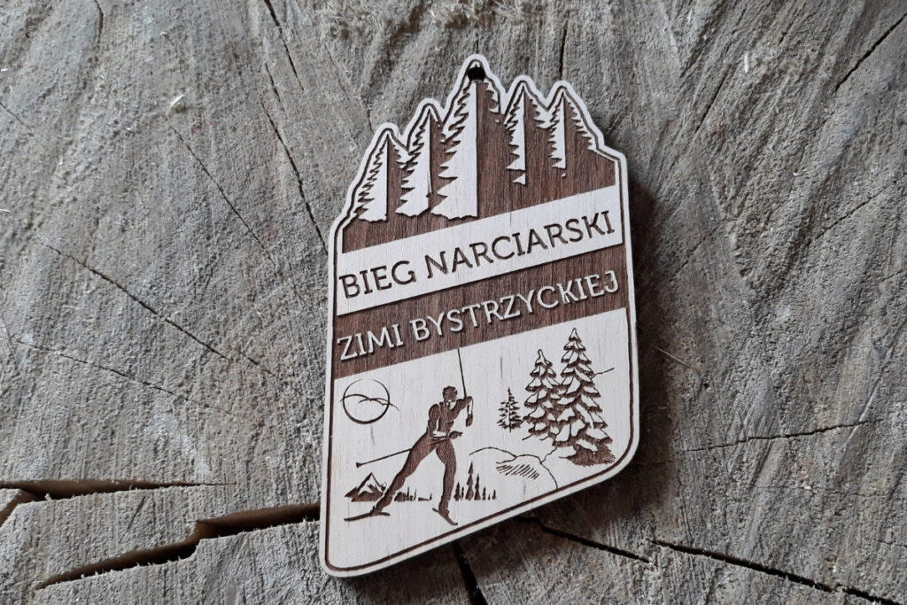 Drewniany medal na „Bieg Narciarski Ziemi Bystrzyckiej”, z precyzyjnym grawerem narciarza biegowego na tle ośnieżonych szczytów górskich, wykonany z jasnej brzozy o nieskazitelnie czystych krawędziach.