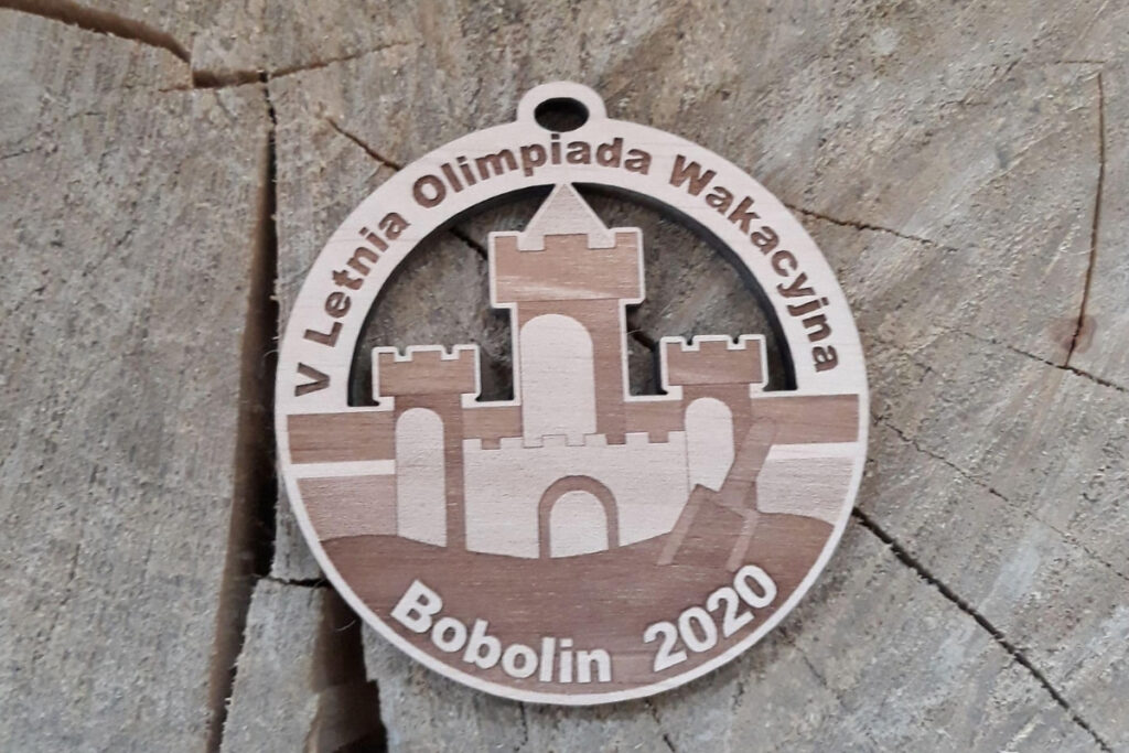 Ażurowy drewniany medal na „Letnią Olimpiadę Wakacyjną” z precyzyjnie wyciętym motywem bramy zamkowej, oraz napisem okolicznościowym, wykonany z jasnej brzozy o krystalicznie czystych krawędziach.