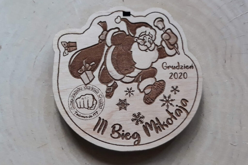 Drewniany medal na „III Bieg Mikołaja”, z radosnym grawerem biegnącego Świętego Mikołaja w czapce z pomponem, otoczony motywami płatków śniegu i nazwą zawodów, wykonany z jasnej brzozy bez przypaleń.