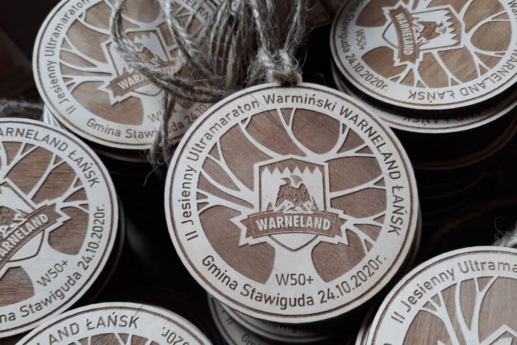 Okrągły drewniany medal na „Warneland – Jesienny Ultramaraton Warmiński”, z grawerem logotypu biegu, motywem jesiennych gałęzi drzewa oraz dystansem, wykonany z czystej brzozy bez śladów dymu i osmaleń.