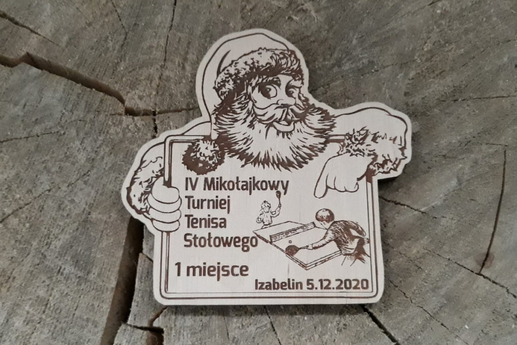 Drewniany medal na „Mikołajkowy Turniej Tenisa Stołowego”, w kształcie mikołaja trzymającego tablicę, z grawerem 2 osób grających w ping-ponga oraz świątecznej czapki Mikołaja, wykonany z czystej brzozy bez osmaleń.