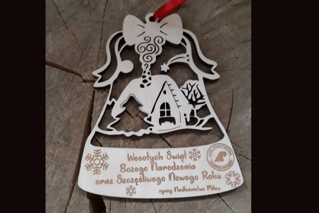 Ażurowa drewniana bombka w kształcie dzwonka z wyciętym domkiem pośród śniegu oraz wygrawerowanymi personalizowanymi życzeniami świątecznymi, wykonana z jasnej brzozy bez śladów osmaleń.