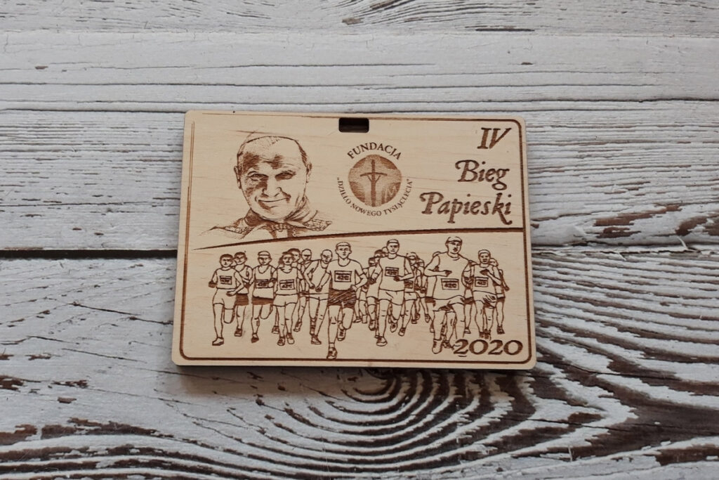 Prostokątny drewniany medal na „Bieg Papieski”, z precyzyjnym grawerem wizerunku św. Jana Pawła II, sylwetkami biegaczy oraz nazwą wydarzenia, wykonany z czystej brzozy bez osmaleń, grubość medalu 6 mm.